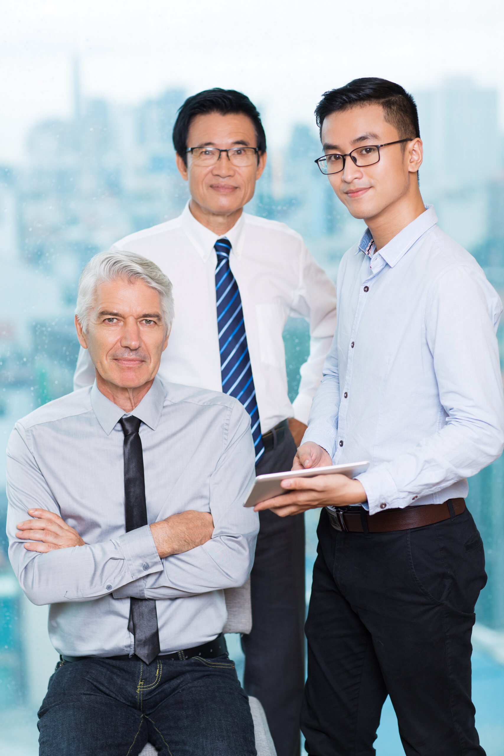 Portrait of three confident businessmen. Senior Caucasian man with crossed arms sitting on chair, young manager holding tablet, Asian business expert looking at camera. Team concept